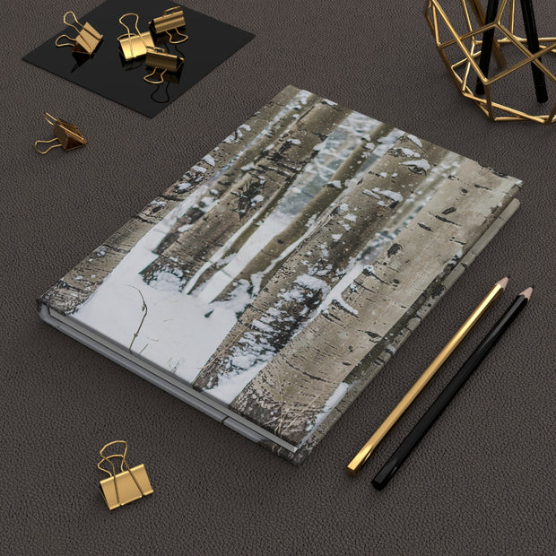 A hardcover notebook with a winter birch tree print lies on a dark textured surface, styled with gold and black binder clips, two pencils, and a gold geometric decor object.