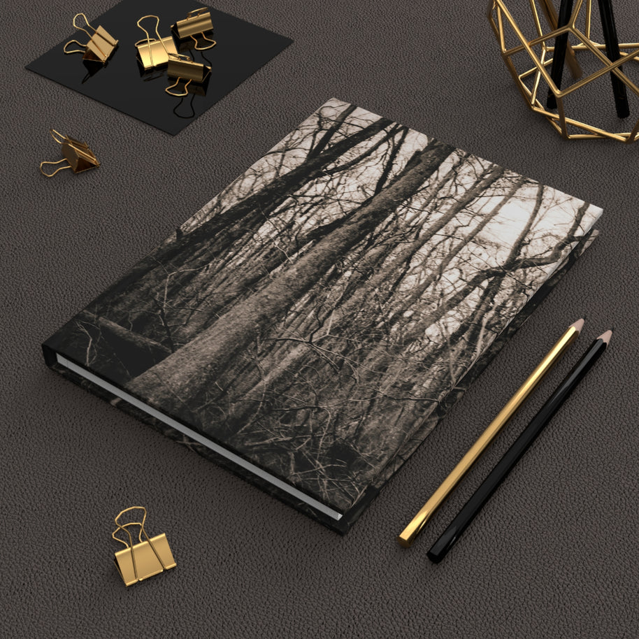 A hardcover notebook with a black and white forest tree branch pattern on the cover, styled on a dark textured surface with gold and black office accessories.