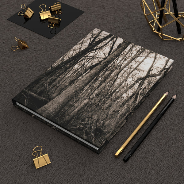 A hardcover notebook with a black and white forest tree branch pattern on the cover, styled on a dark textured surface with gold and black office accessories.