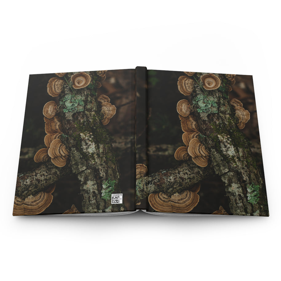 An open hardcover book or journal displayed flat against a white background, featuring a photographic cover design of brown shelf mushrooms growing on tree bark with green moss.