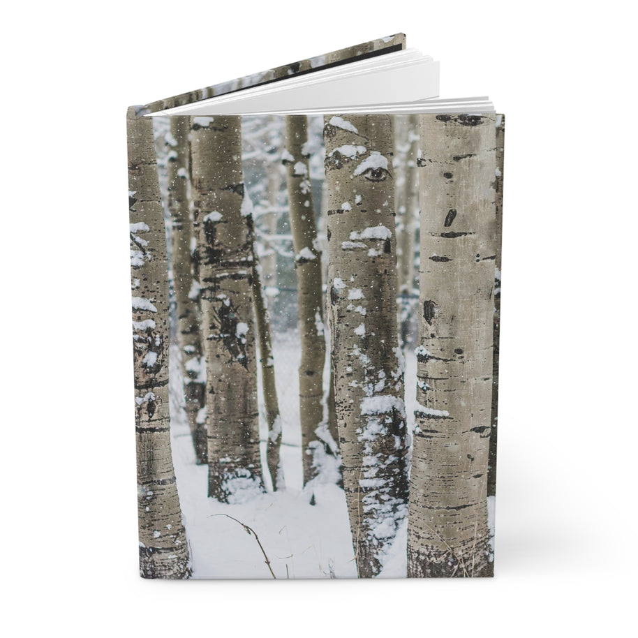 A hardcover notebook or journal standing upright, featuring a winter forest cover with birch tree trunks and snow on the cover, photographed on a clean white background.