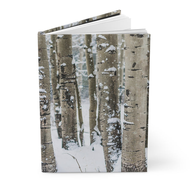A hardcover notebook or journal standing upright, featuring a winter forest cover with birch tree trunks and snow on the cover, photographed on a clean white background.
