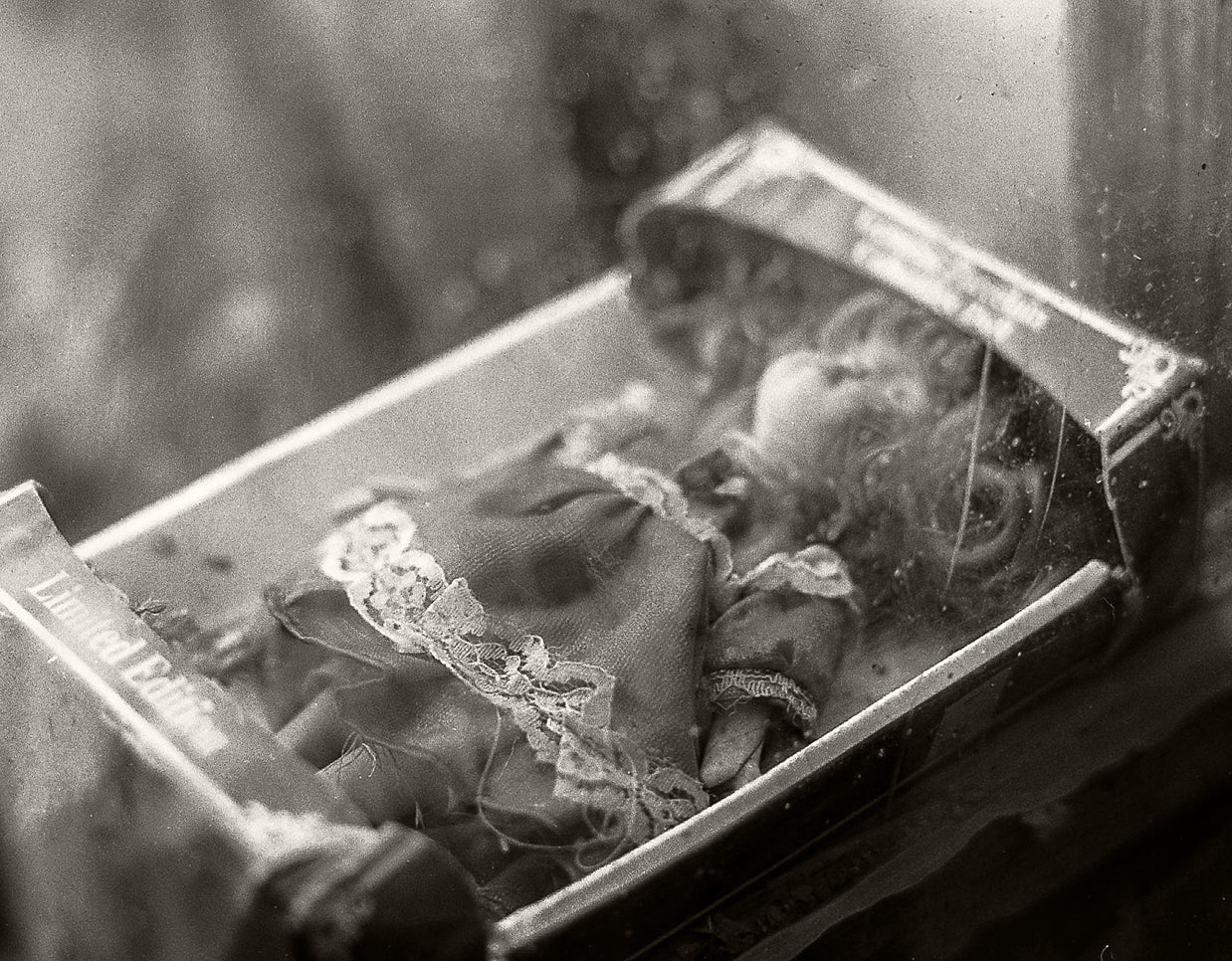 A vintage black and white photograph of a small doll or figurine inside an open book or box, with a blurred, atmospheric background.