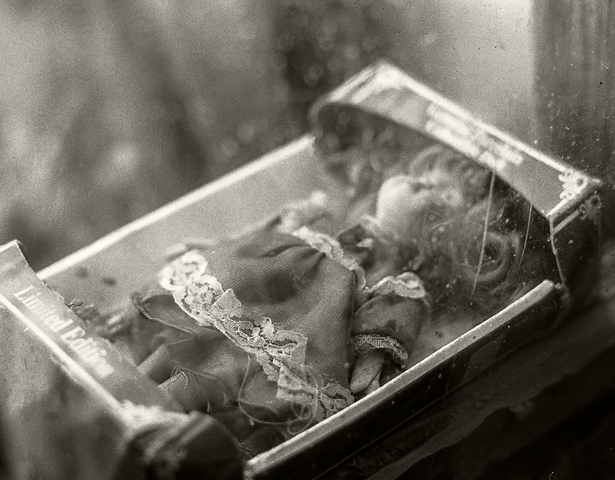 A vintage black and white photograph of a small doll or figurine inside an open book or box, with a blurred, atmospheric background.