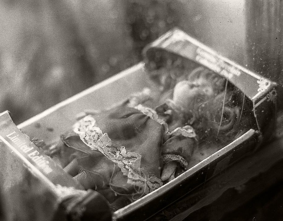 A vintage black and white photograph of a small doll or figurine inside an open book or box, with a blurred, atmospheric background.