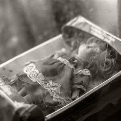 A vintage black and white photograph of a small doll or figurine inside an open book or box, with a blurred, atmospheric background.