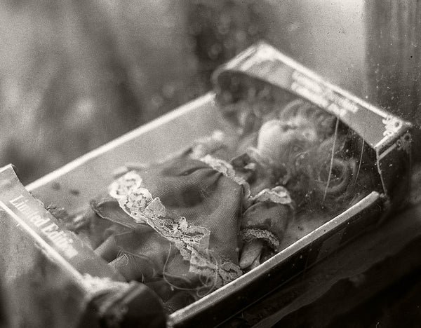 A vintage black and white photograph of a small doll or figurine inside an open book or box, with a blurred, atmospheric background.