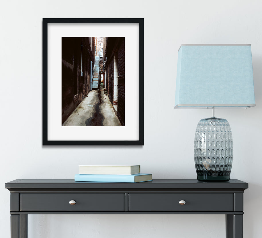 Black-framed alabama photo print of grunge alley birmingham alabama with dark walls and wet pavement above gray console table